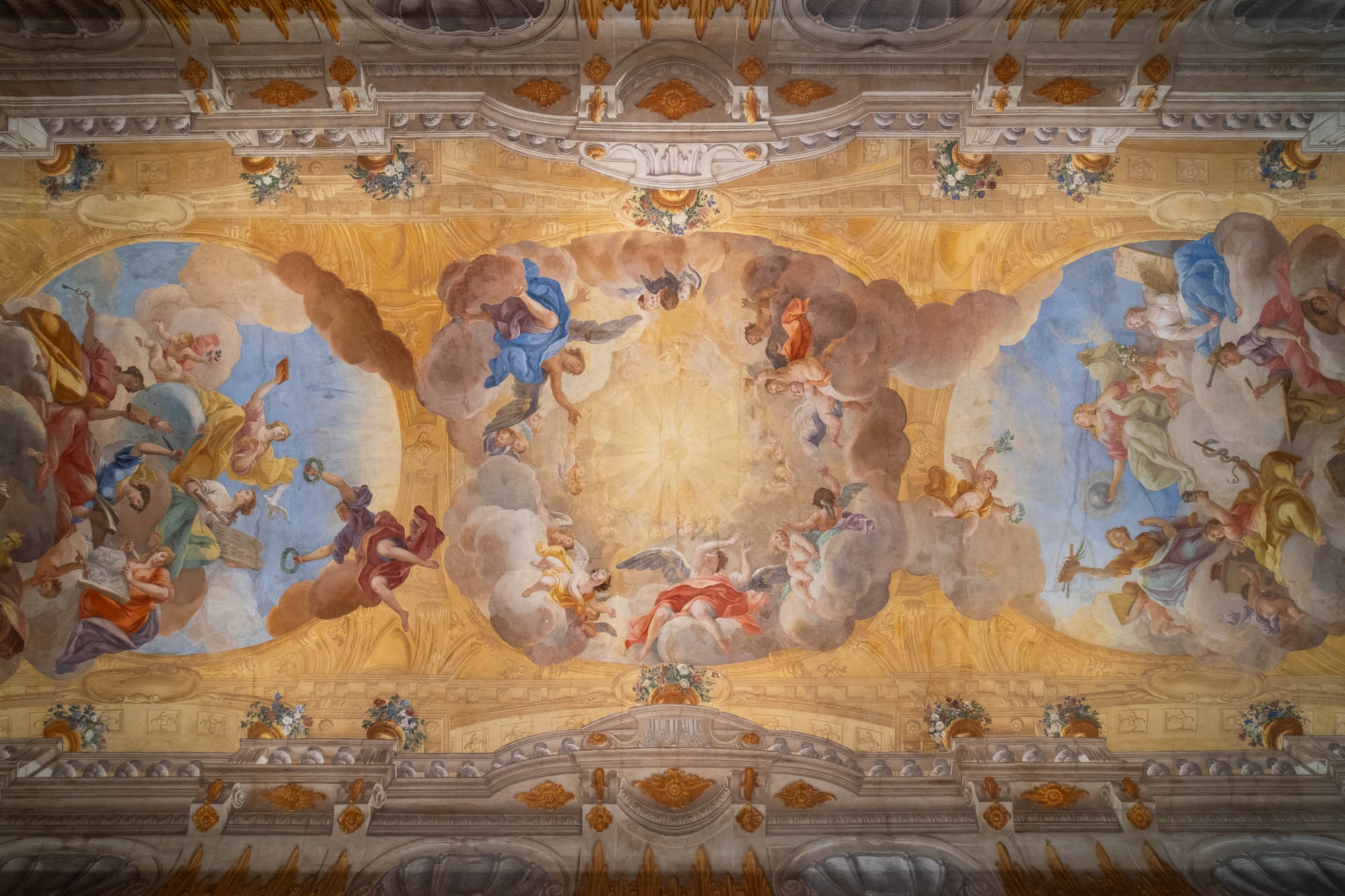 A detailed ceiling fresco featuring numerous figures in flowing garments, clouds, and architectural elements, with a central sunlit area surrounded by classical motifs and ornamental designs.