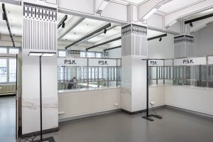 A spacious, modern room with high ceilings, white walls, and decorative columns. Several empty service counters labeled “P.S.K.” line the area; one person sits alone at a desk in the background.