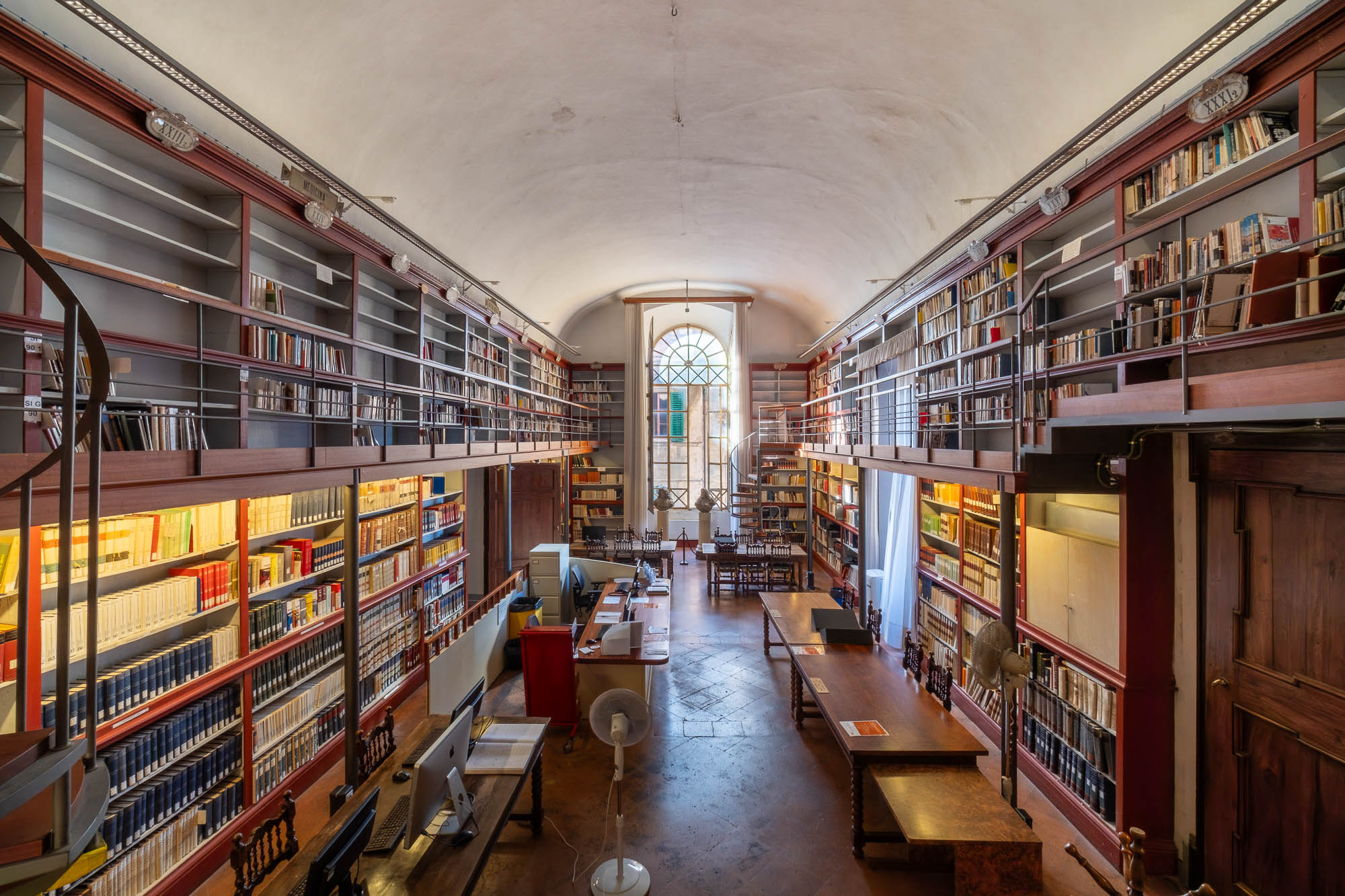A large, well-lit library room with tall bookshelves on two levels, filled with books. There are desks, chairs, and a few computers in the center, and a large arched window at the far end lets in natural light.