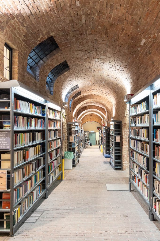 A long, arched hallway in a library with brick ceilings and walls, lined on both sides with tall bookshelves filled with books. A person sits at a desk at the very end,