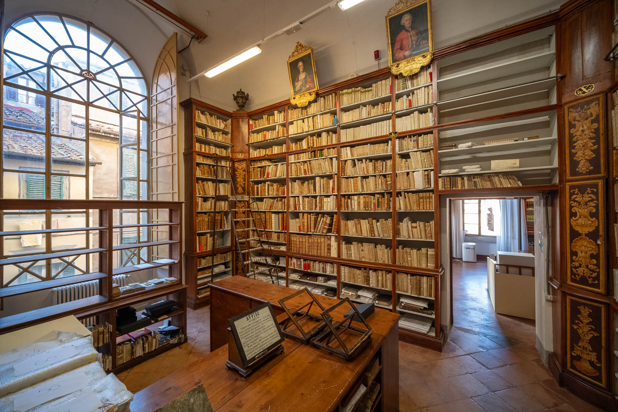 A historic library room with tall bookshelves filled with books, a wooden desk with display stands, a large arched window, a wheeled ladder, paintings above the shelves, and an open doorway leading to another room.