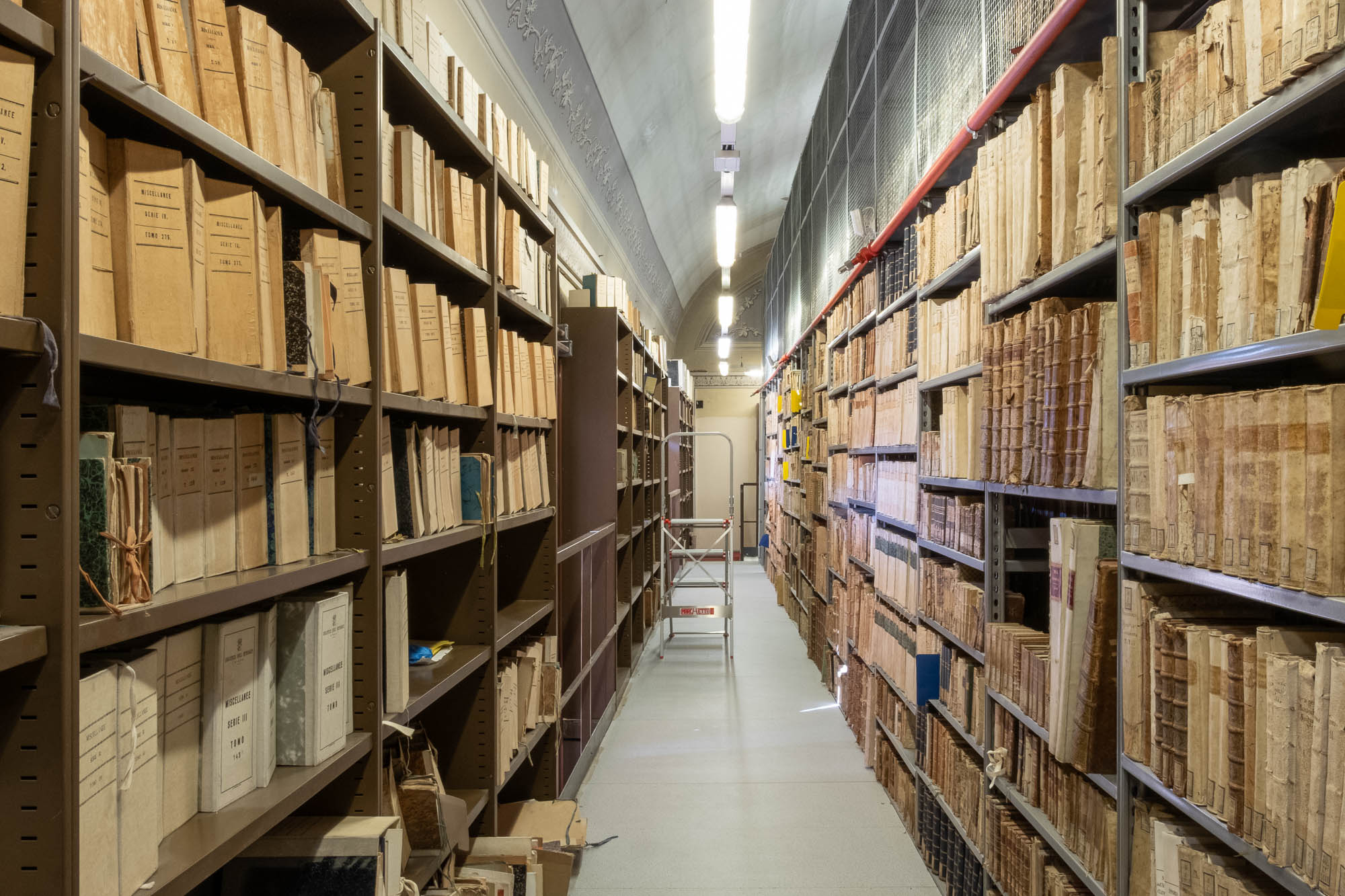 Narrow aisle between tall shelves filled with old books and documents in brown, worn covers; a step ladder is positioned at the end of the aisle under bright ceiling lights.