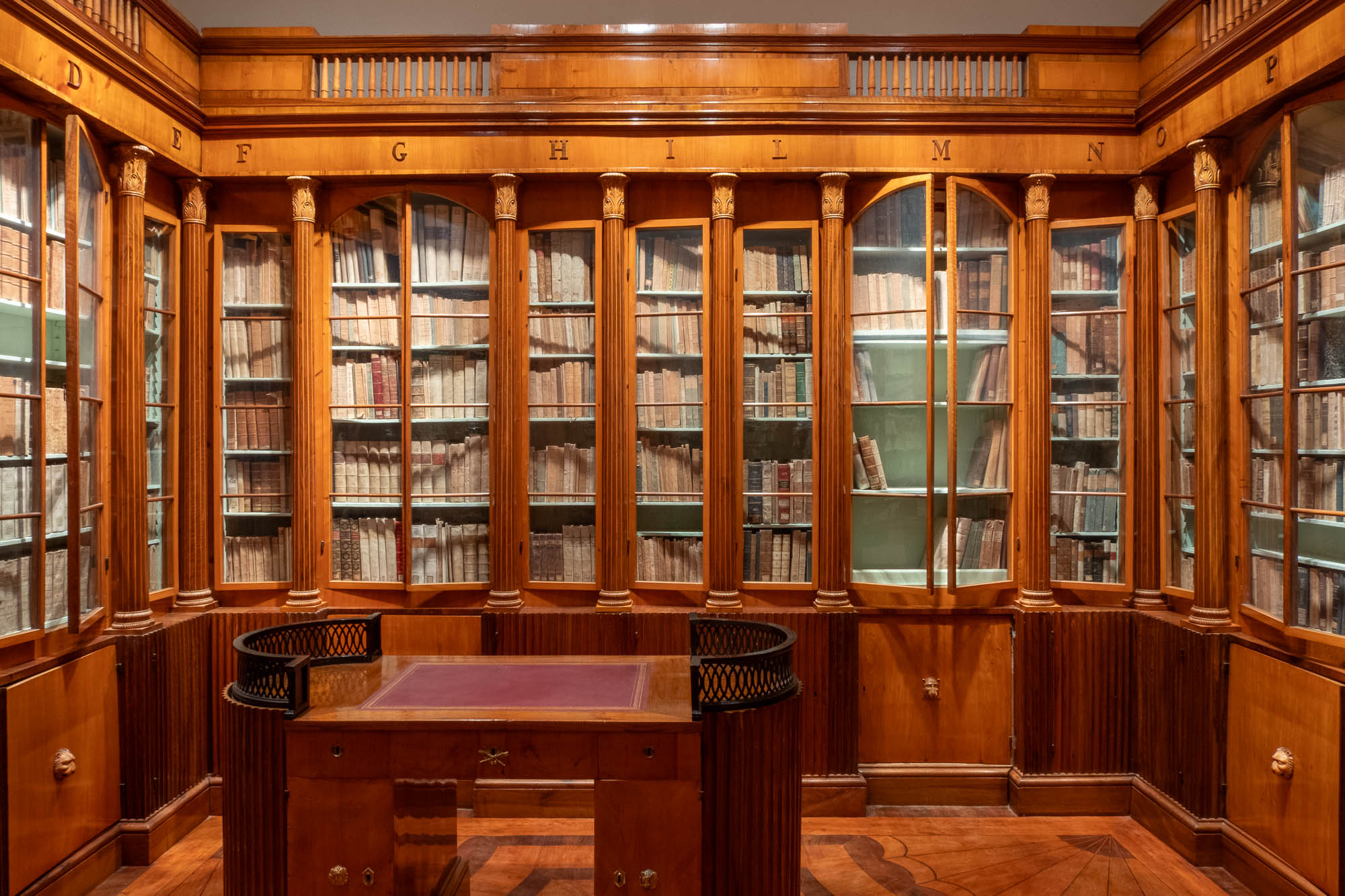 A wooden library room with glass-fronted bookshelves filled with old books and mark with letters at the top. A wooden desk stands in the middle.