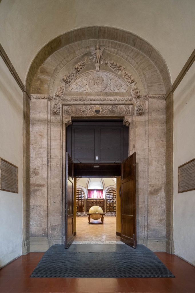 Open wooden doors lead to a room with arched stonework and ornate carvings above the entrance. Inside, a large globe stands in the center surrounded by chairs, with red curtains in the background.