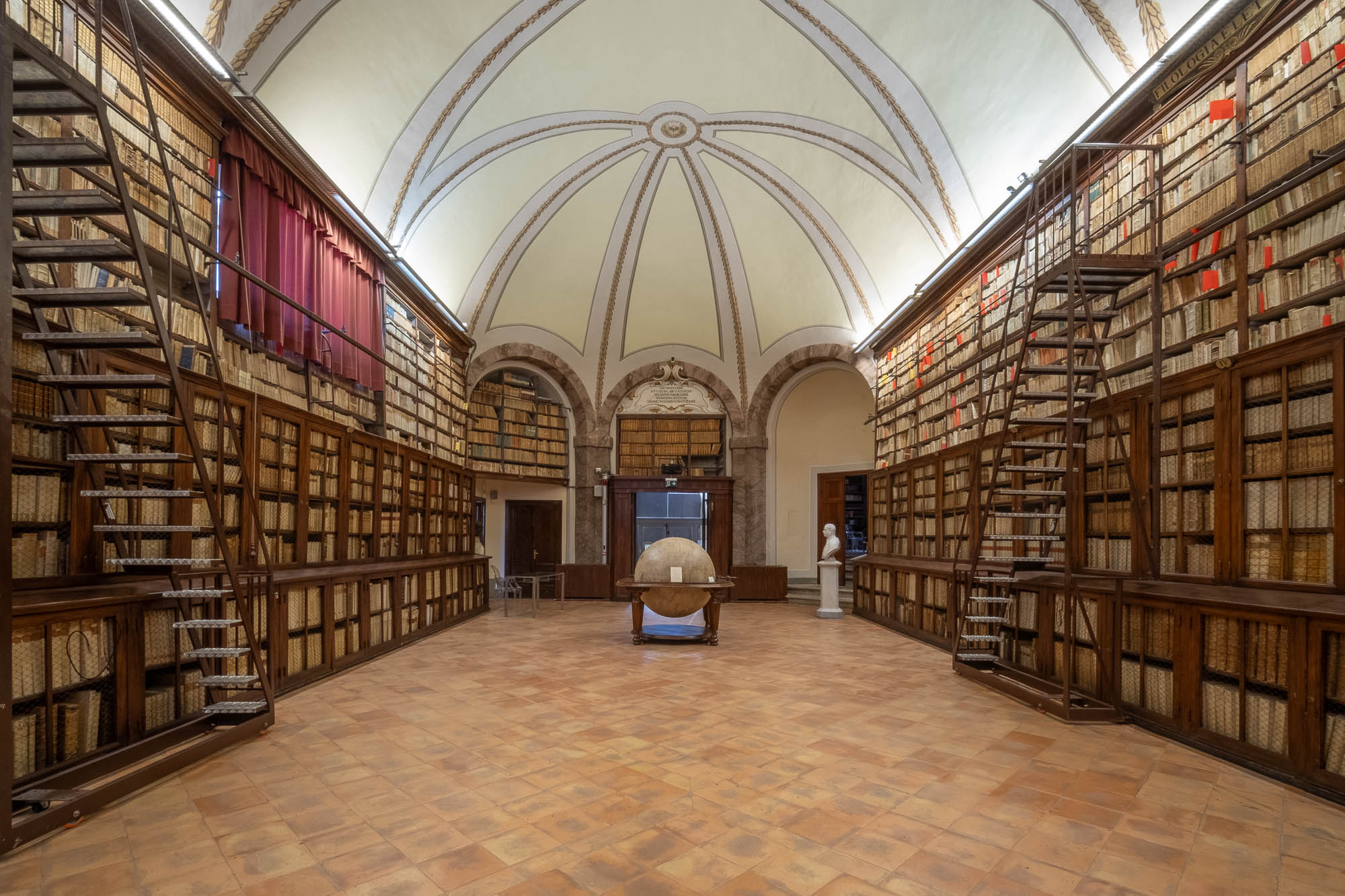 A spacious library room with high arched ceilings, tall bookshelves filled with old books, two rolling ladders, and a large globe in the center of the tiled floor; a white statue stands in the background.