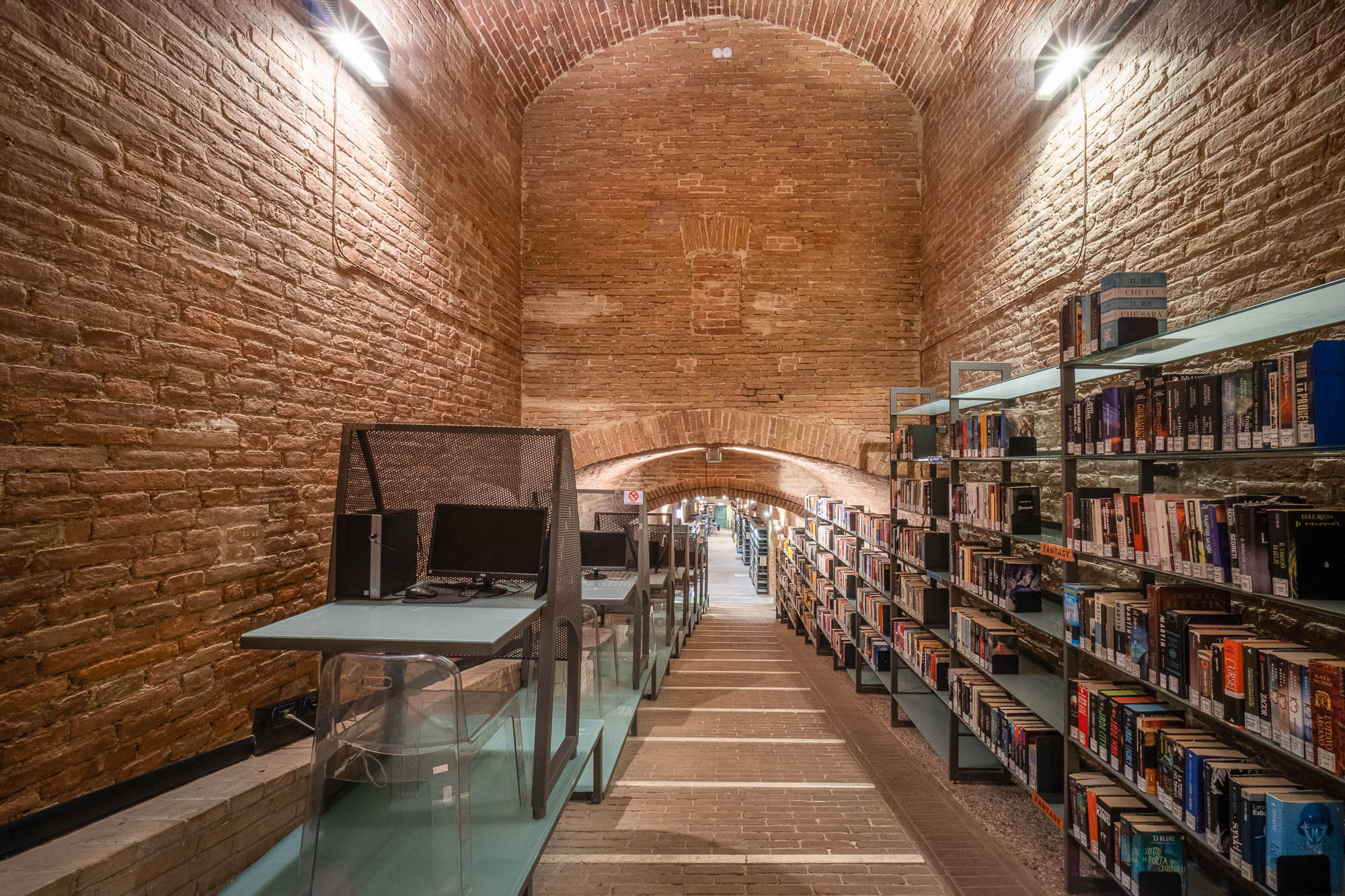 A library with brick walls and vaulted ceiling, empty computer desks on the left, and bookshelves filled with books on the right; bright lighting illuminates the space.