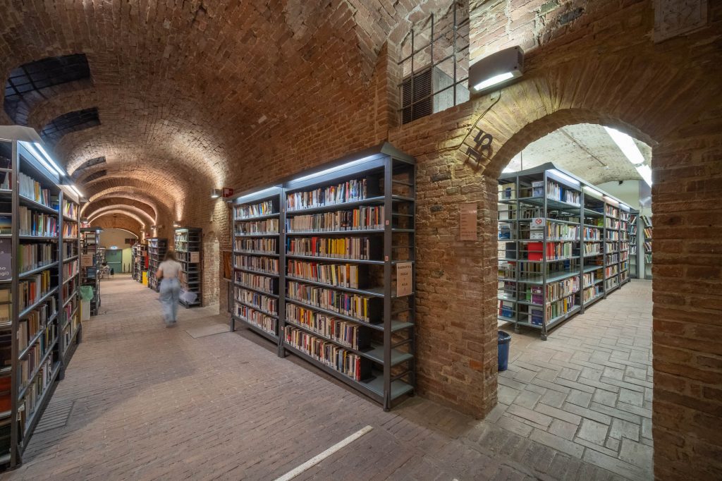 A person walks through a brick-arched library with bookshelves filled with books on both sides and another section visible through an arched doorway.