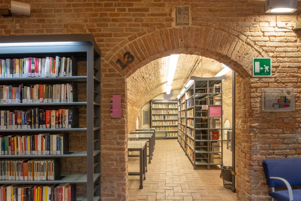 A brick archway labeled 13 leads to bookshelves and study desks in a well-lit library. Shelves are filled with books, and an emergency exit sign is mounted on the wall to the right of the arch.