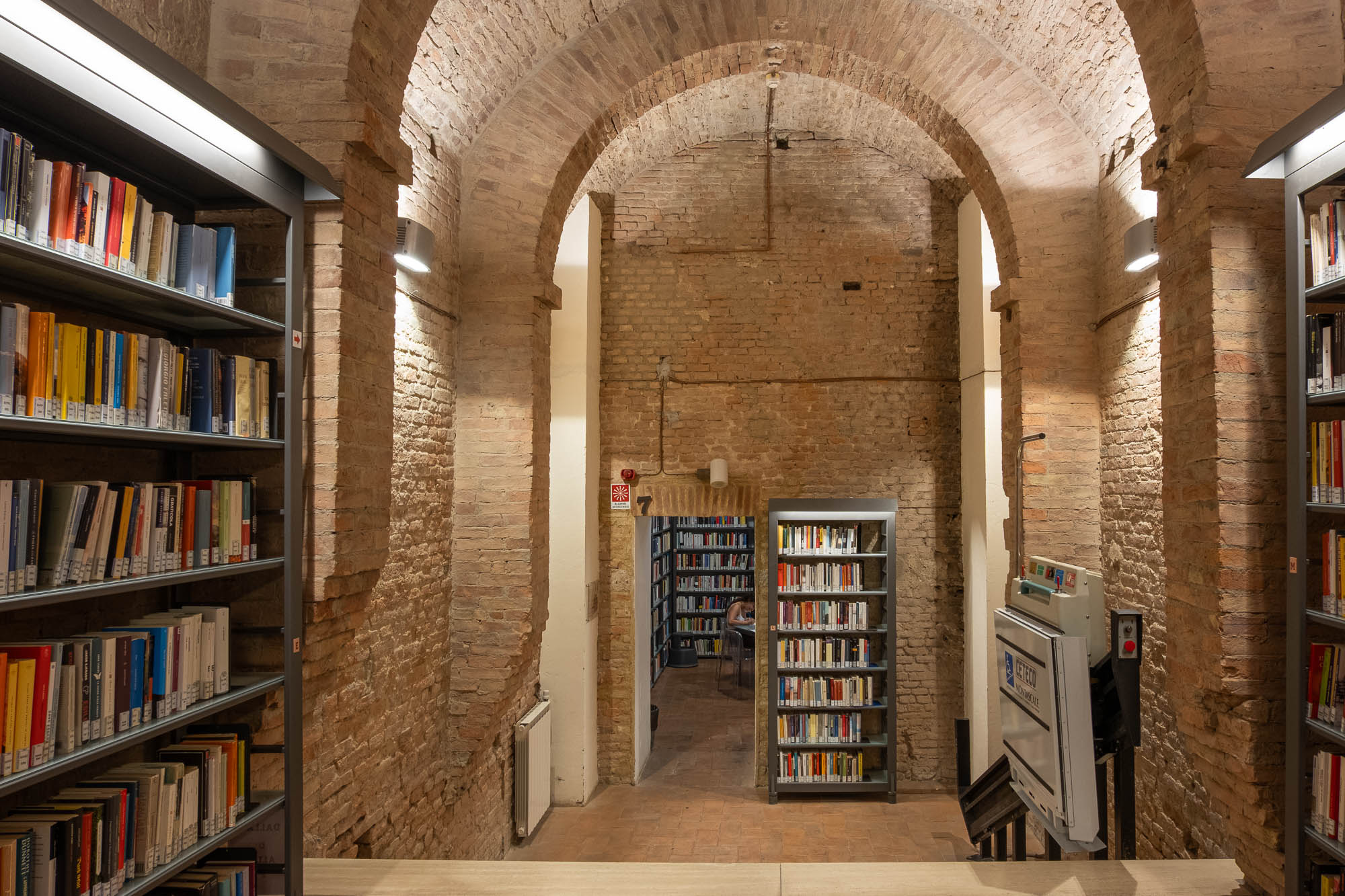 A brick-walled library interior with arched ceilings, rows of bookshelves filled with books, and a stairlift attached to a stone staircase leading to a lower level with more bookshelves.