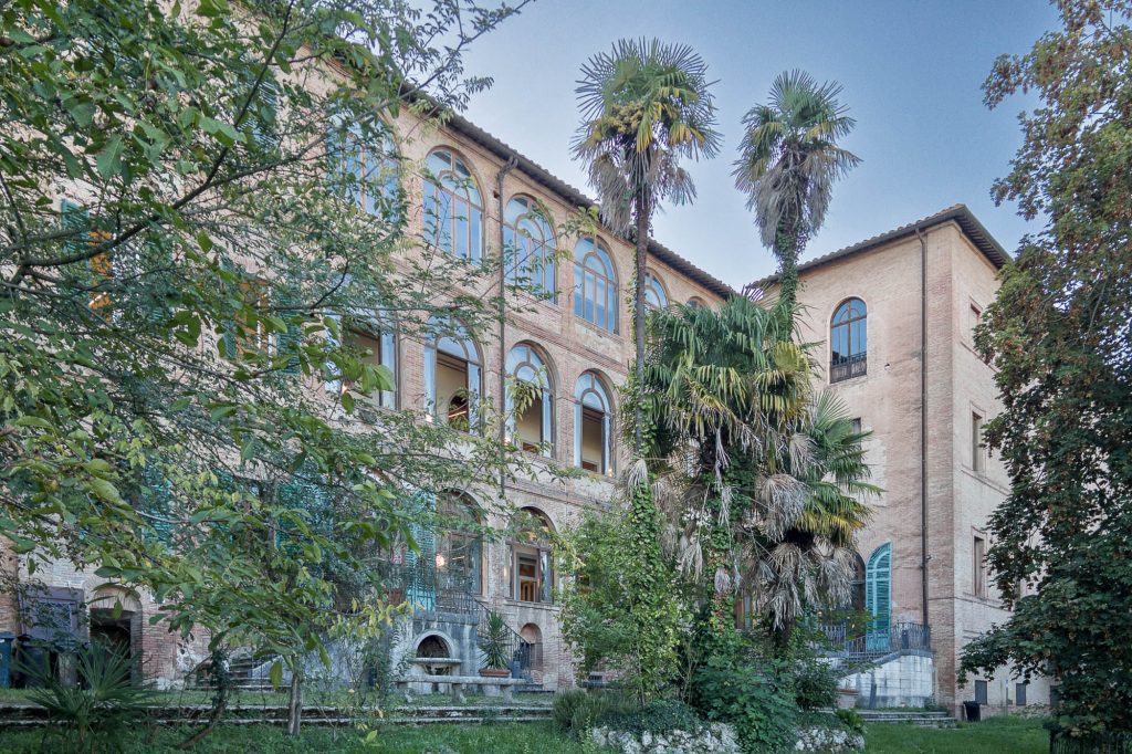 A large brick building with arched windows and multiple stories is surrounded by tall palm trees and dense greenery under a clear sky.