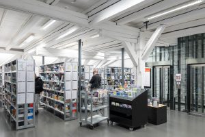 A modern library interior with white beams, tall shelves filled with books, two people browsing, and metal carts nearby. An elevator with glass doors is visible on the right side.