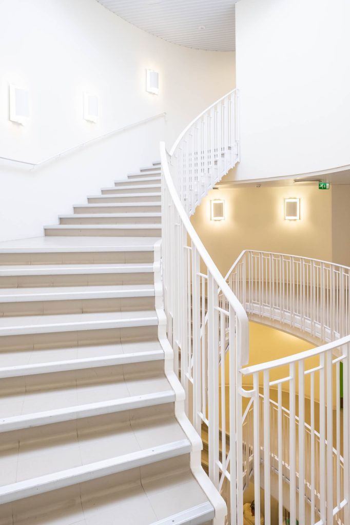 A clean, modern staircase with white railings curves upward in a brightly lit interior space. The walls are white, with several rectangular wall lights mounted along the staircase.