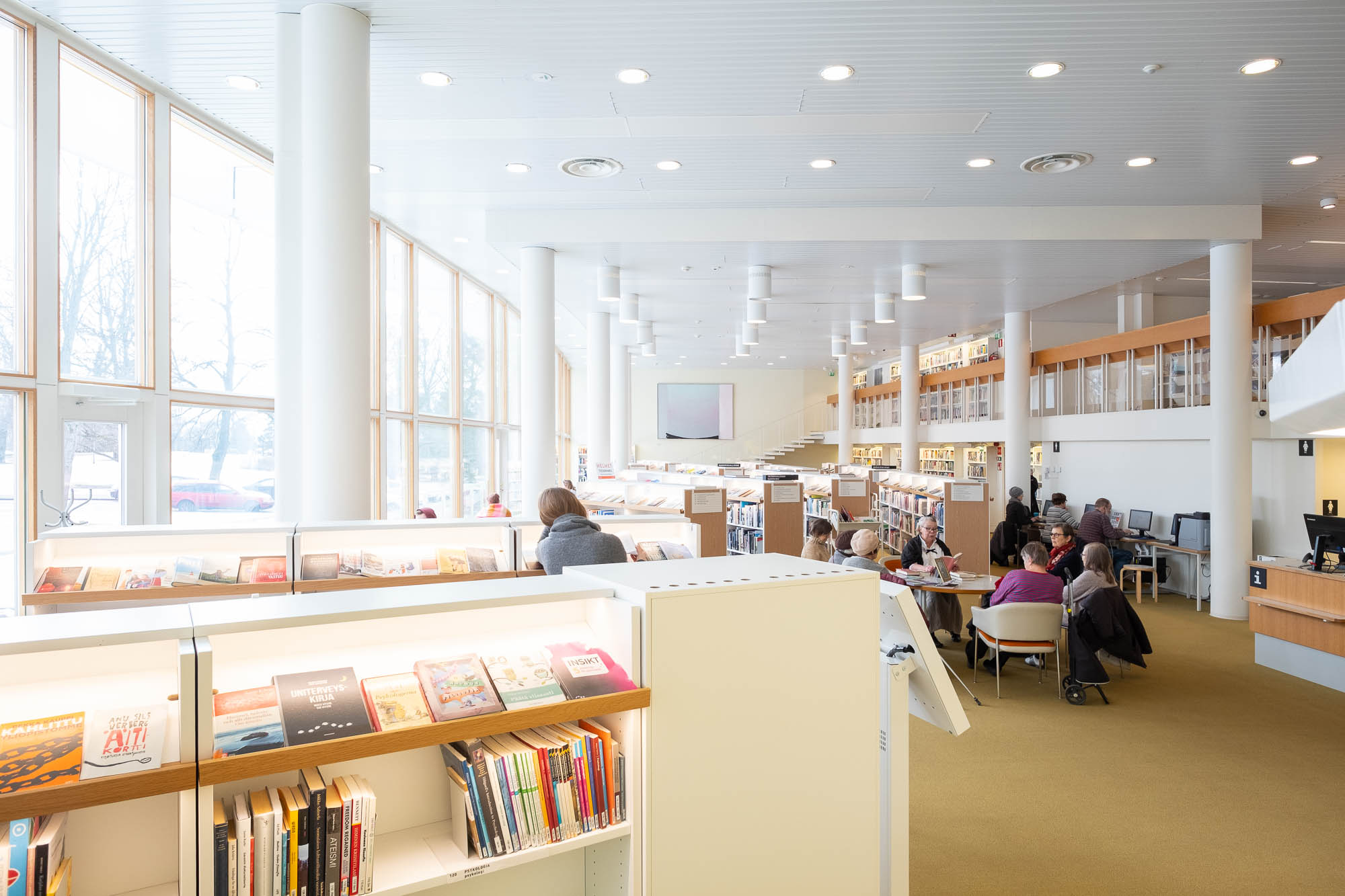 Large, modern library interior with tall white columns, rows of bookshelves filled with books, high ceilings, and large windows letting in natural light. Several people are sitting and reading or browsing books.