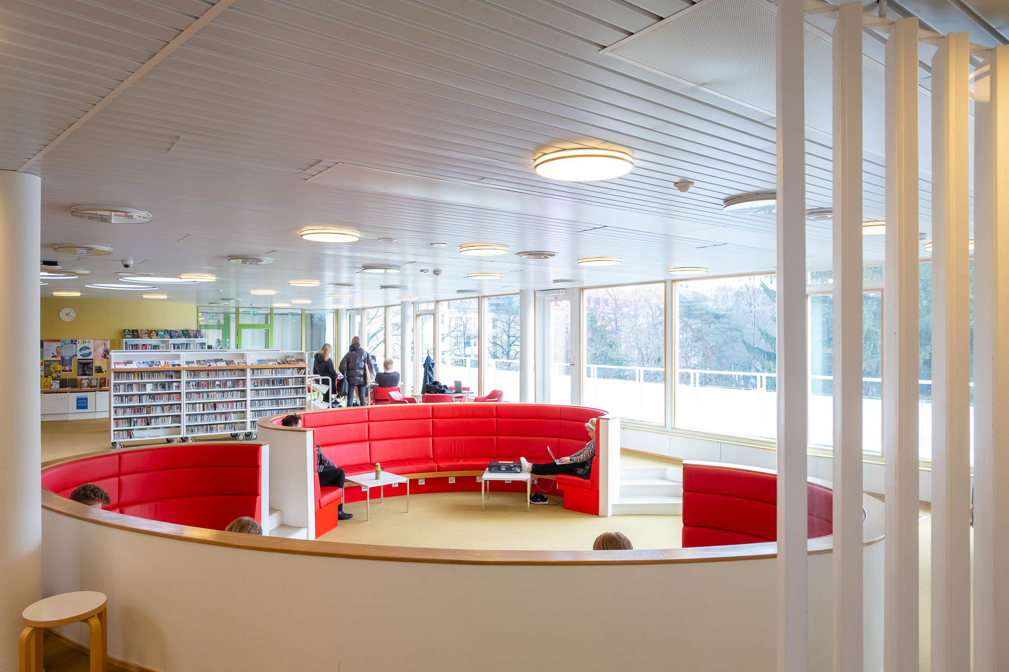 A modern library interior with curved red seating in the center, several people reading or talking, bookshelves in the background, and large windows letting in natural light.