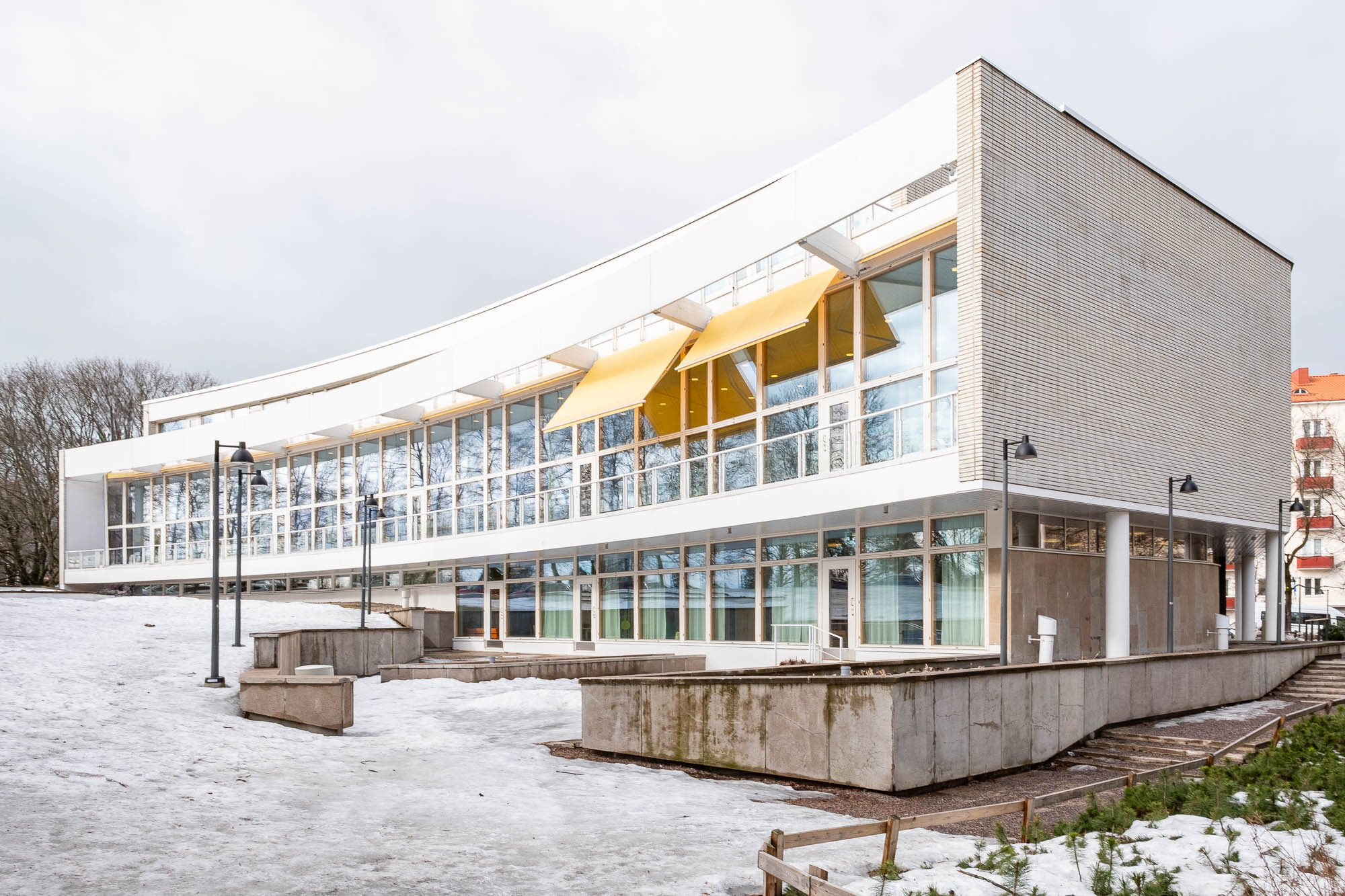 A modern two-story building with large glass windows and yellow awnings, surrounded by snow and concrete walkways, with leafless trees and a few other buildings in the background.