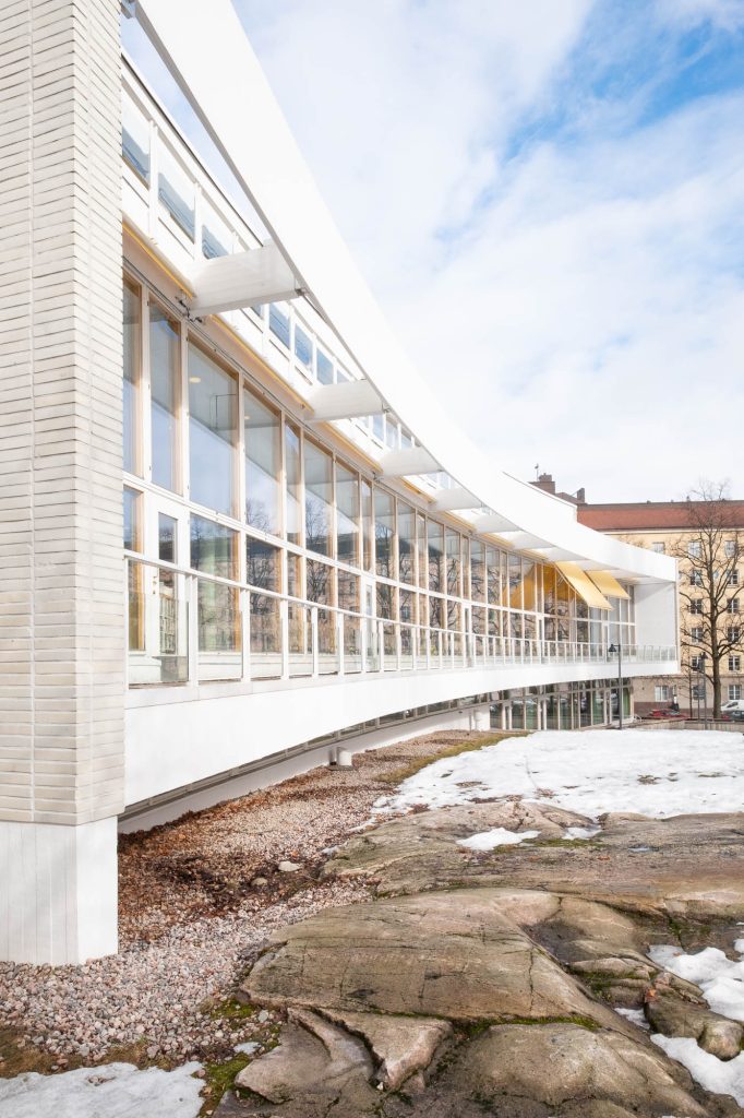A modern building with large glass windows and a curved white roof, elevated above a rocky ground with patches of snow. The sky is partly cloudy and other buildings are visible in the background.