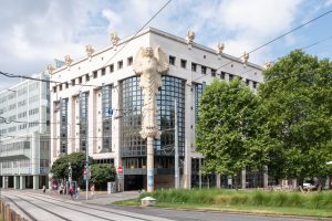 A modern, multi-story building with large windows and distinctive sculpted figures along the roofline and corner representing owls. Trees, tram lines, and pedestrians are visible in the foreground under a partly cloudy sky.