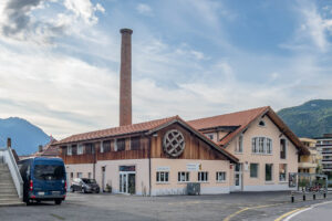A light-colored building with a red tile roof and a prominent circular wooden feature on the facade stands next to a tall brick chimney. Several cars and a blue van are parked outside, with mountains visible in the background.