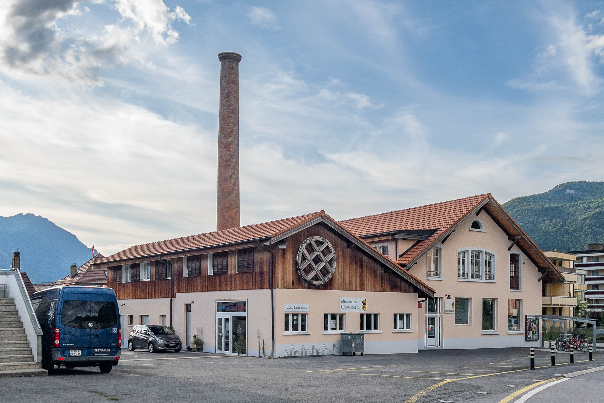 A light-colored building with a red tile roof and a prominent circular wooden feature on the facade stands next to a tall brick chimney. Several cars and a blue van are parked outside, with mountains visible in the background.