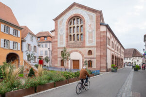 A person rides a bicycle past a large Neo-Romanesque building of red stone, arched windows and a decorative facade that bears the words "Stadtbibliothek - Museum" and "Bibliothèque Humaniste". There is a patch of greenery at the front.