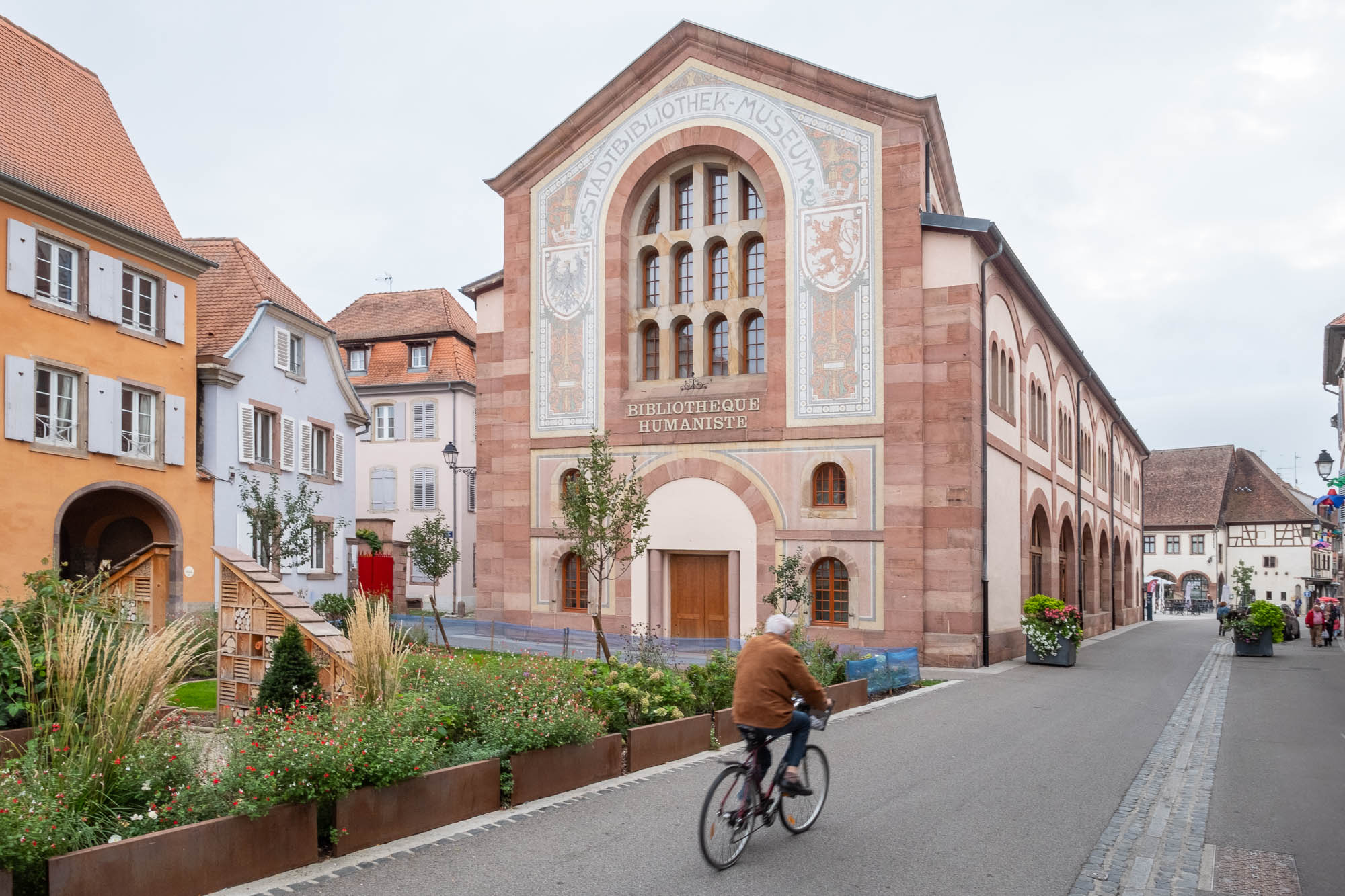 A person rides a bicycle past a large Neo-Romanesque building of red stone, arched windows and a decorative facade that bears the words "Stadtbibliothek - Museum" and "Bibliothèque Humaniste". There is a patch of greenery at the front.