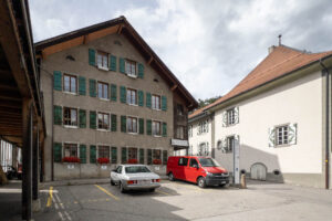 A three-story building with green shutters and flower boxes stands beside a white building with painted window frames. Two vehicles, a white car and a red van, are parked in marked spaces on a paved lot.