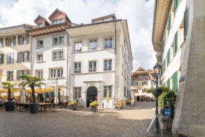 A cobblestone square bordered by white and beige buildings, outdoor cafes with covered tables, potted plants, and a few parked bicycles on a sunny day. People sit at tables and some walk through the square.
