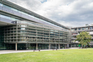 Modern glass and steel building with large windows and metal framing, adjacent to a grassy lawn and small trees, under a partly cloudy sky. Benches and a person on a bike are visible near the building.
