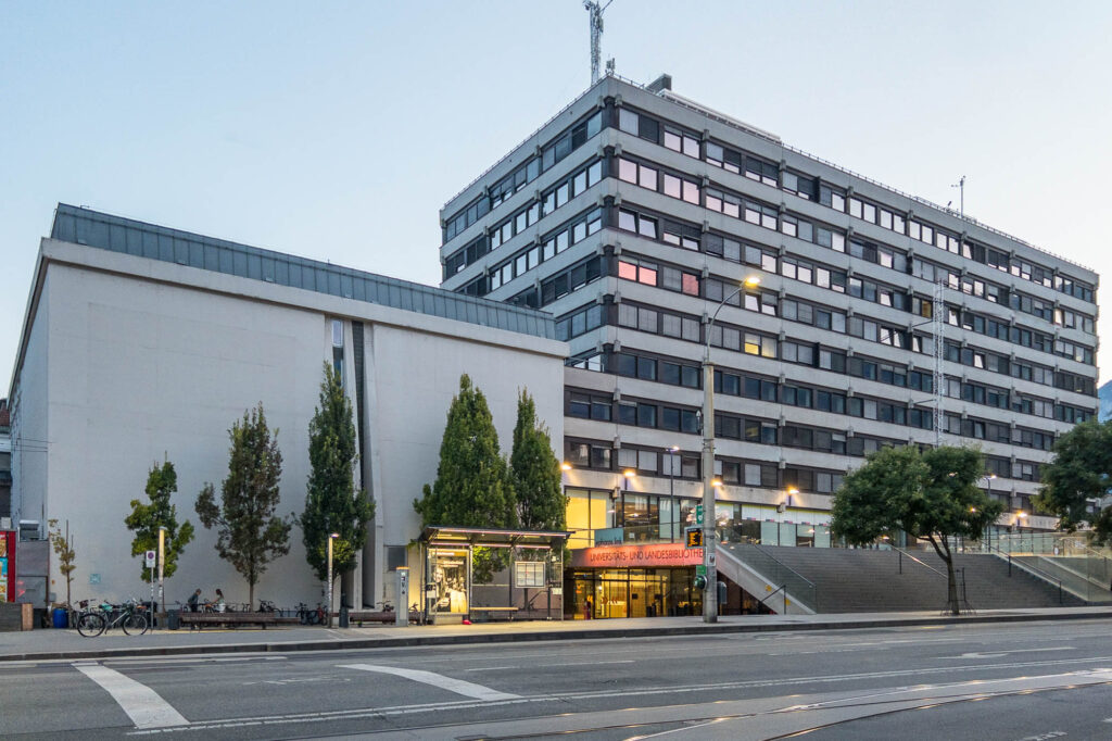 A modern, multi-story office building with many windows stands beside a smaller structure. There are trees, street lamps, bicycles, and a few people near the entrance. A wide stairway leads up to a terrace. Underneath it is the entrance of the Universitäts- und Landesbibliothek, indicated with a glowing sign.