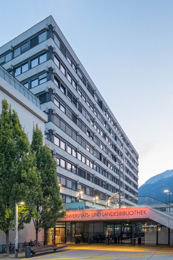 A multi-story modern building with many windows, labeled Universitäts- und Landesbibliothek, stands beside green trees. A mountain is visible in the background under a clear sky.