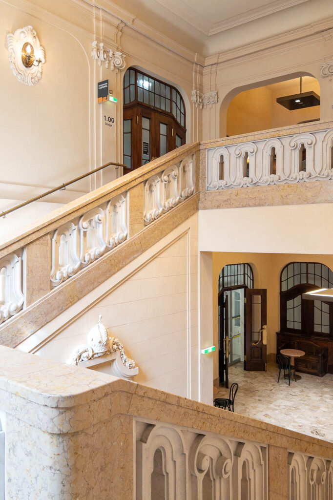 A grand interior staircase with ornate railings and marble details leads to wooden doors and arched windows; tables and chairs are visible in the lower hall. The space is well-lit and decorated in neutral tones.