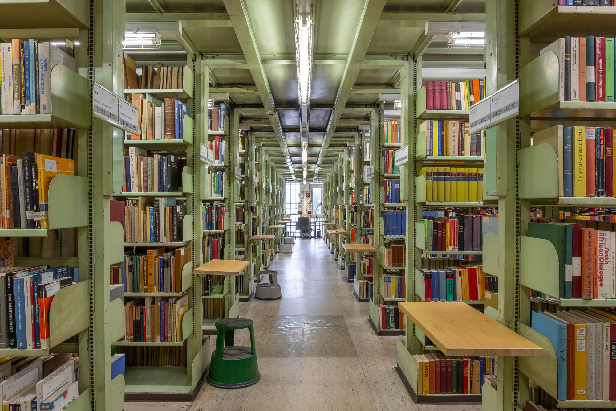 Interior of a book storage block with tall green metal bookshelves filled with books on both sides, small desks attached to the shelves, a step stool on the floor, and a window with natural light at the far end.