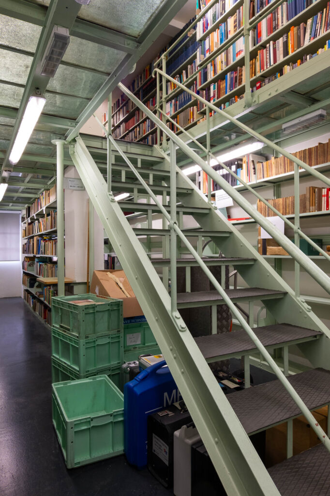 A metal staircase in a library leads to an upper level with bookshelves filled with books; green plastic crates and cardboard boxes are stacked below the stairs.