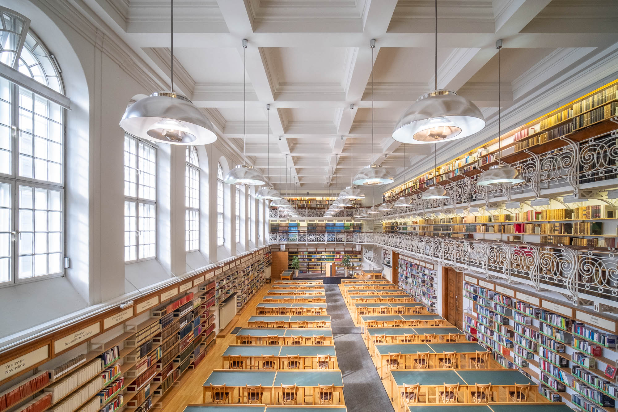 A spacious library with long wooden tables and chairs arranged in rows, tall bookshelves filled with books on two levels, large windows letting in natural light, and two levels of mezzanines with ornate railings. There is a spiral staircase at the back.