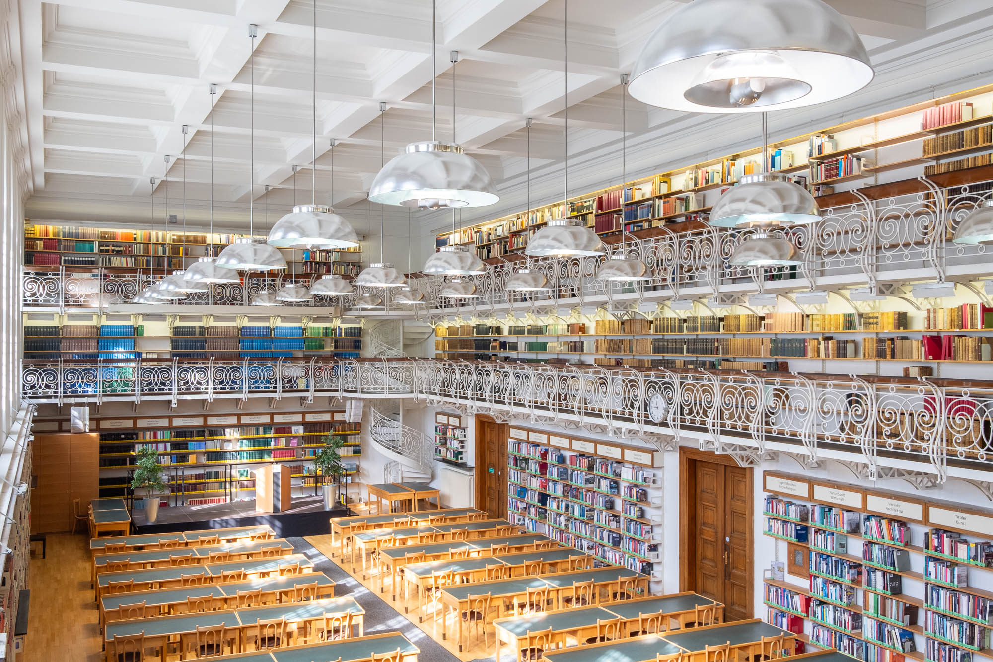 A spacious library with long wooden tables and chairs arranged in rows, tall bookshelves filled with books on two levels, large windows letting in natural light, and two levels of mezzanines with ornate railings. There is a spiral staircase at the back.