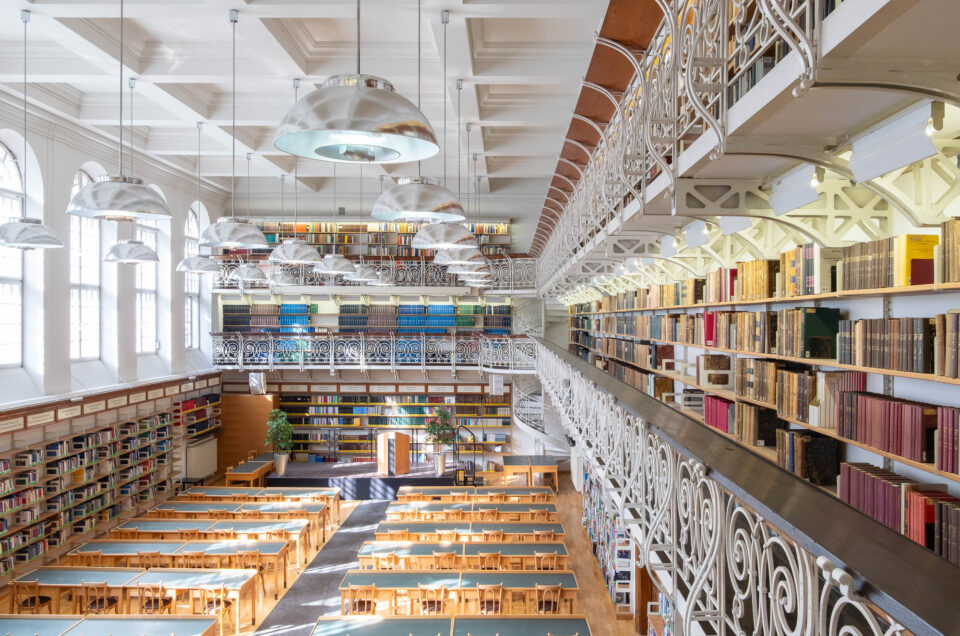 A spacious library with long wooden tables and chairs arranged in rows, tall bookshelves filled with books on two levels, large windows letting in natural light, and two levels of mezzanines with ornate railings. There is a spiral staircase at the back.