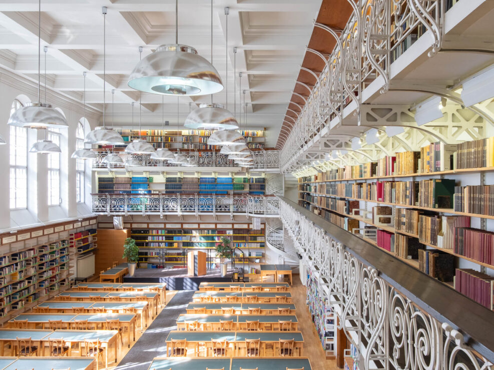 A spacious library with long wooden tables and chairs arranged in rows, tall bookshelves filled with books on two levels, large windows letting in natural light, and two levels of mezzanines with ornate railings. There is a spiral staircase at the back.
