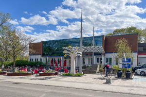 A modern building with large glass windows and red patio umbrellas sits behind a bike rack. A cyclist rides by, and a white metal sculpture stands in the foreground. Trees and partly cloudy sky are visible.