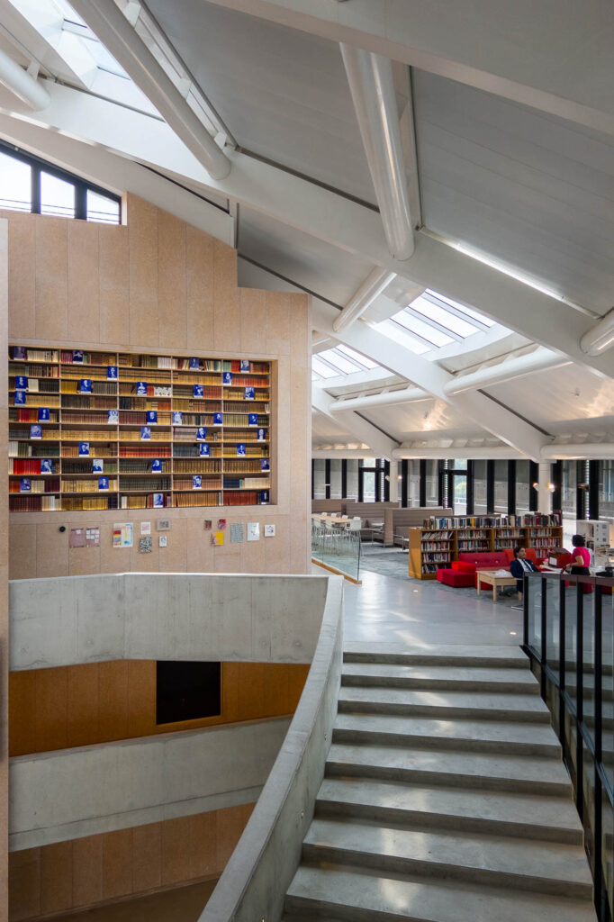 A modern library interior featuring a large, multi-story wall of books, a concrete staircase, and a reading area with red sofas and skylights.