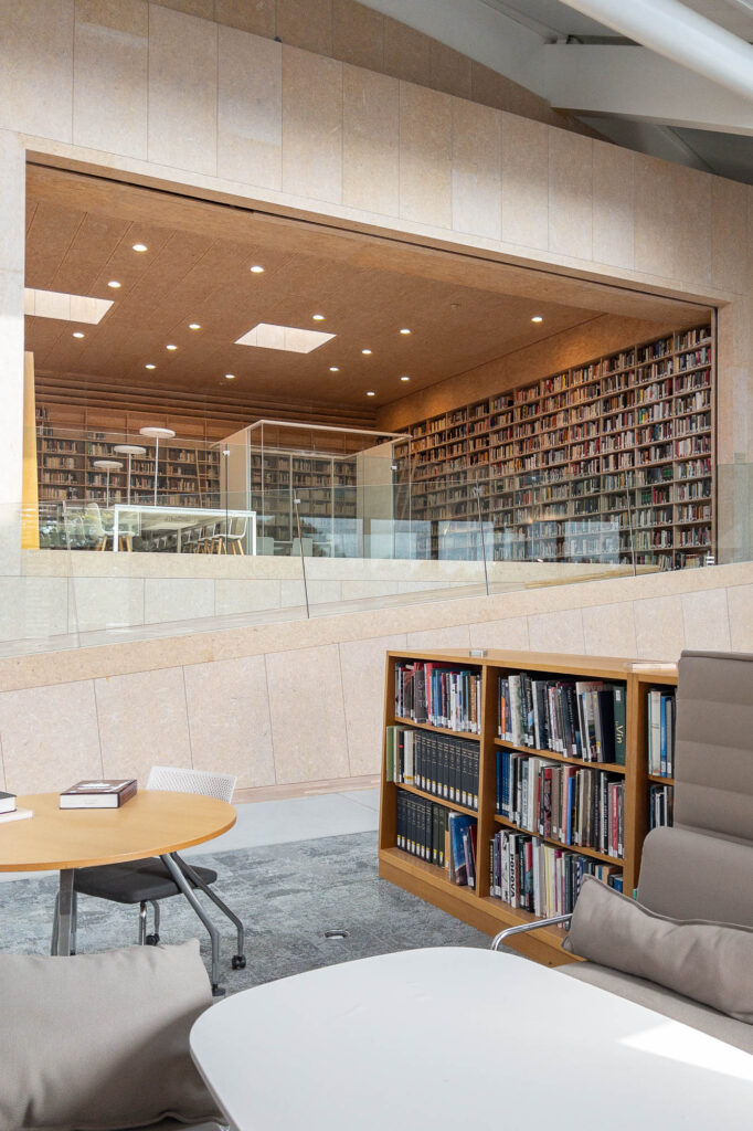 A modern library interior featuring a large, glass-enclosed reading room filled with floor-to-ceiling bookshelves, overlooking a seating area with tables and a bookshelf.