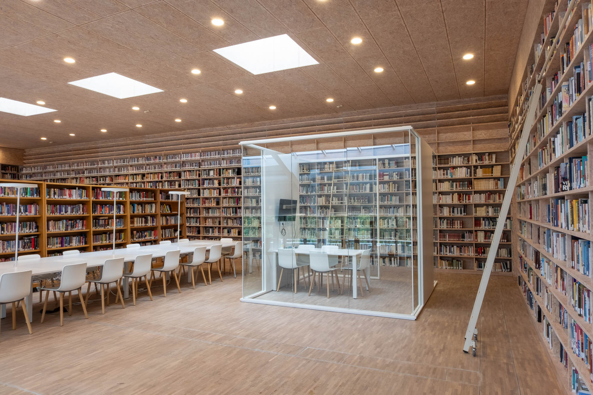 A spacious library with floor-to-ceiling bookshelves, a long reading table with white chairs, and a glass-enclosed study room in the center.