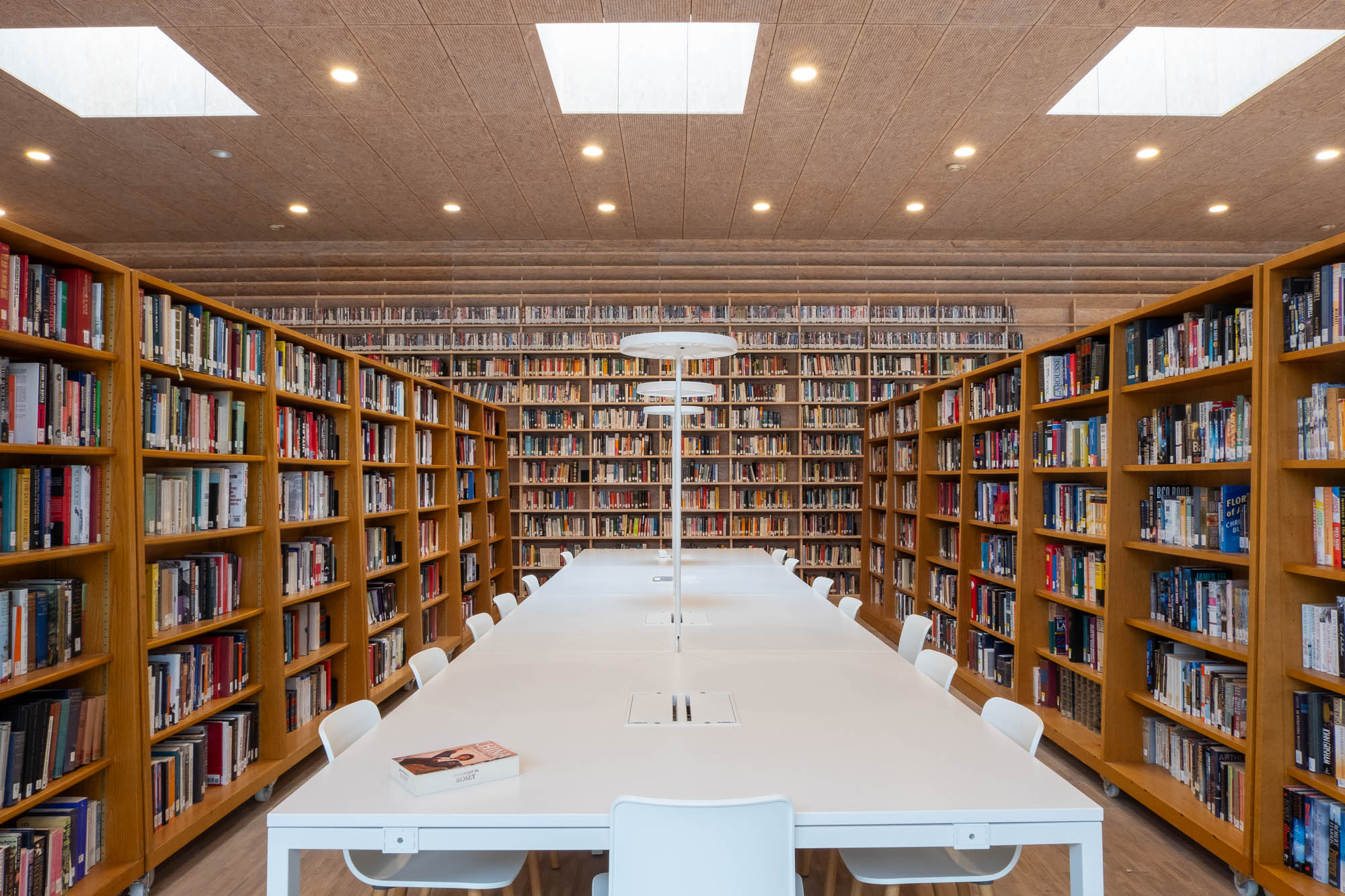 A long white table with chairs sits in the center of a library aisle surrounded by tall wooden bookshelves filled with books.