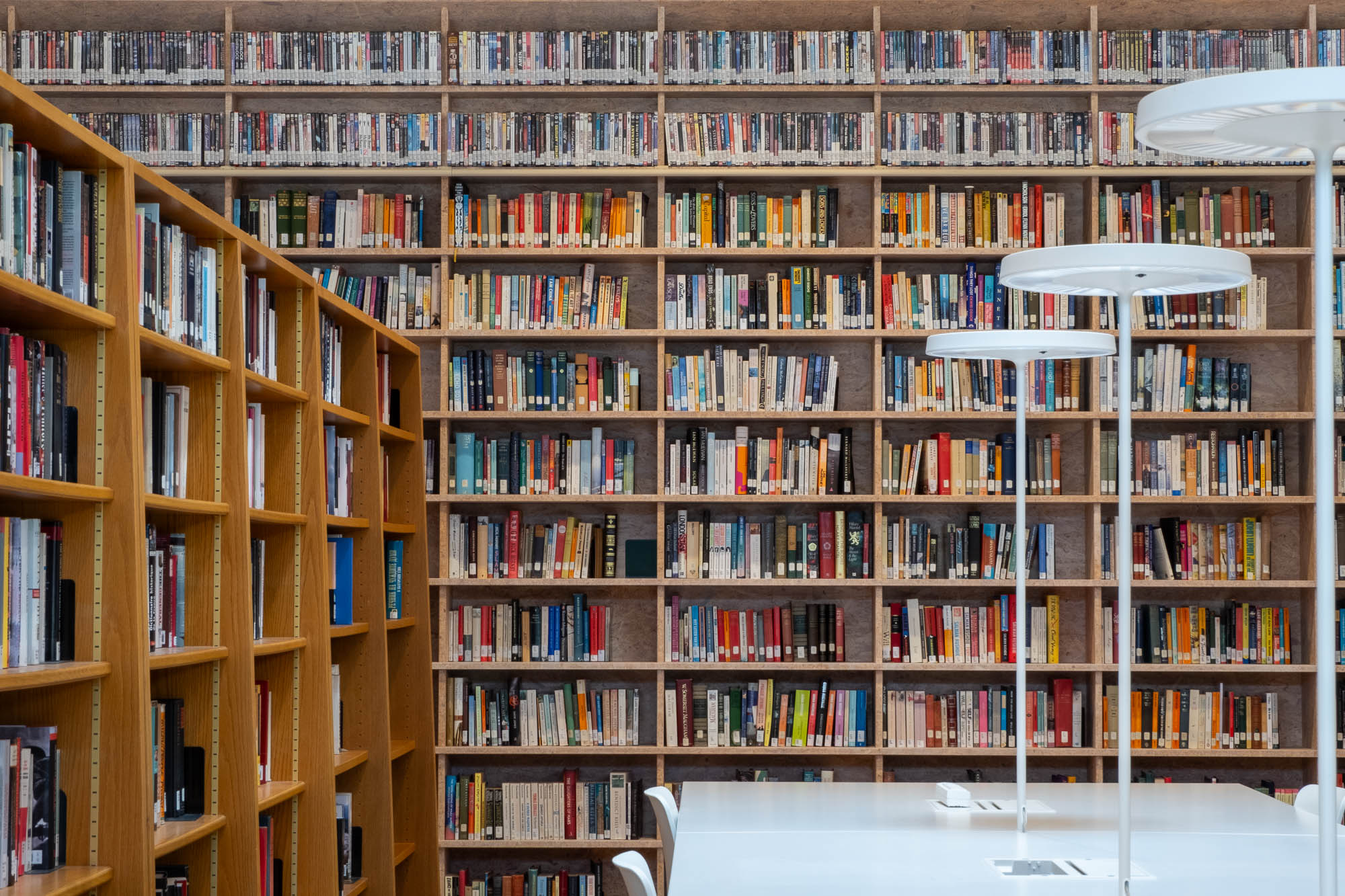 Tall wooden bookshelves filled with rows of books in a library, with a white study table and modern lamps in the foreground.
