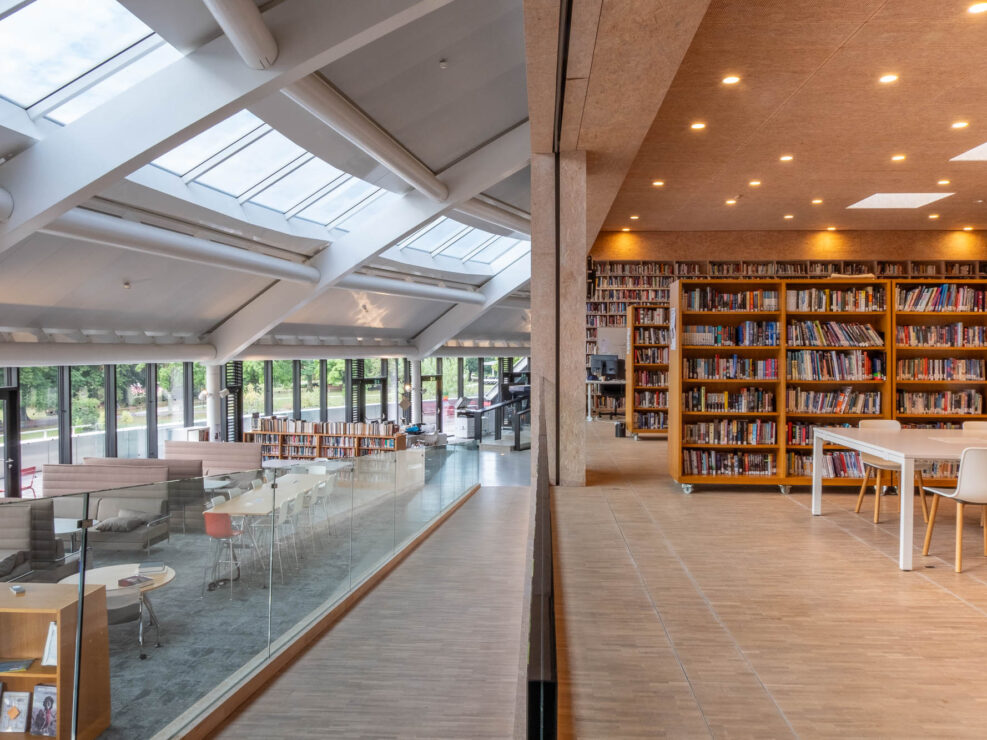 A split-view interior of a modern library, showing a bright, sunlit seating area with skylights on the left and rows of floor-to-ceiling bookshelves with tables on the right.