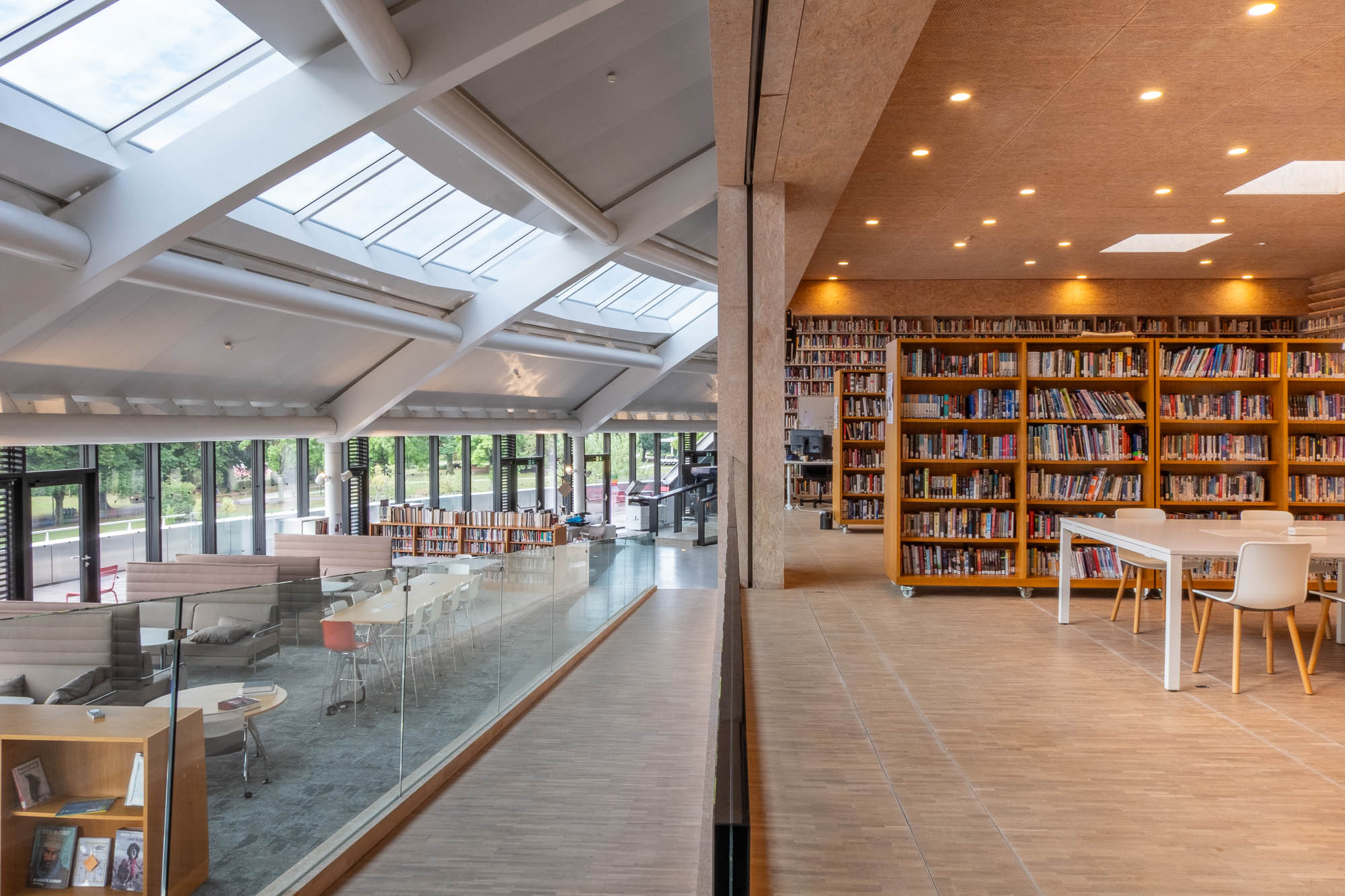 A split-view interior of a modern library, showing a bright, sunlit seating area with skylights on the left and rows of floor-to-ceiling bookshelves with tables on the right.