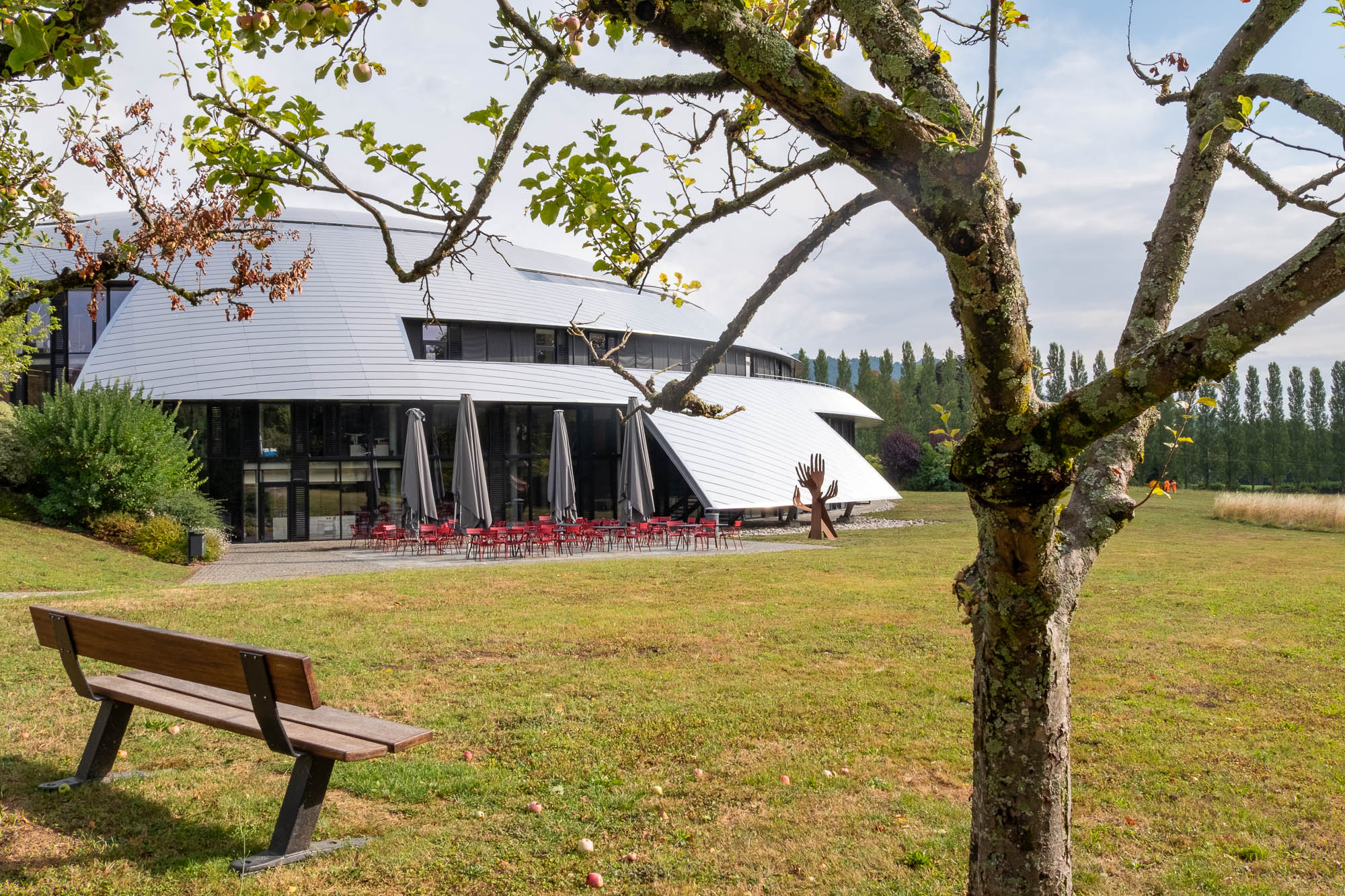 A modern building with a curved, tiered white roof and outdoor patio seating sits in a grassy field with a bench in the foreground.