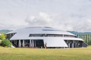 A large, domed modern building with a silver roof and glass walls sits on a grassy lawn.