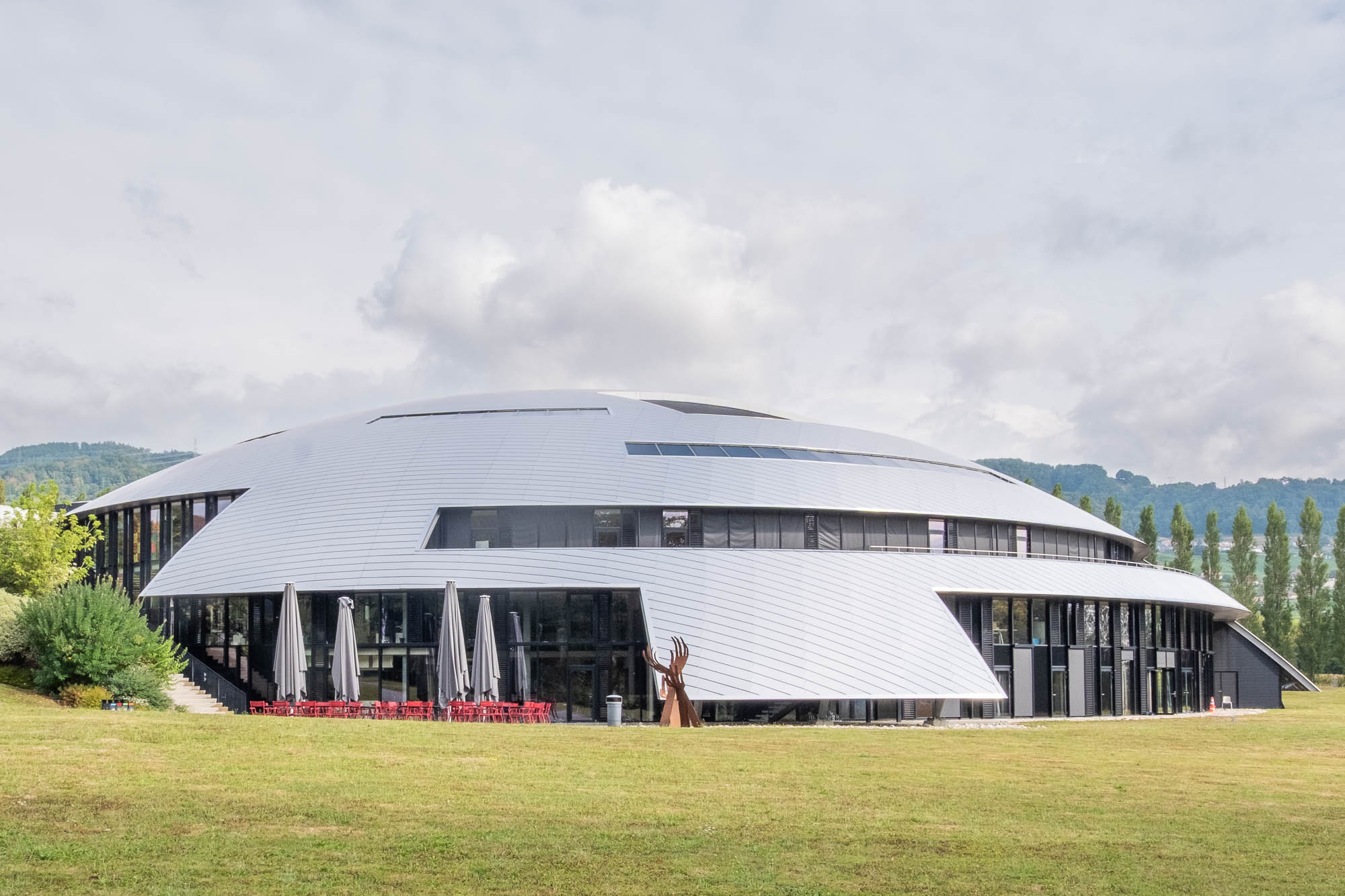A large, domed modern building with a silver roof and glass walls sits on a grassy lawn.